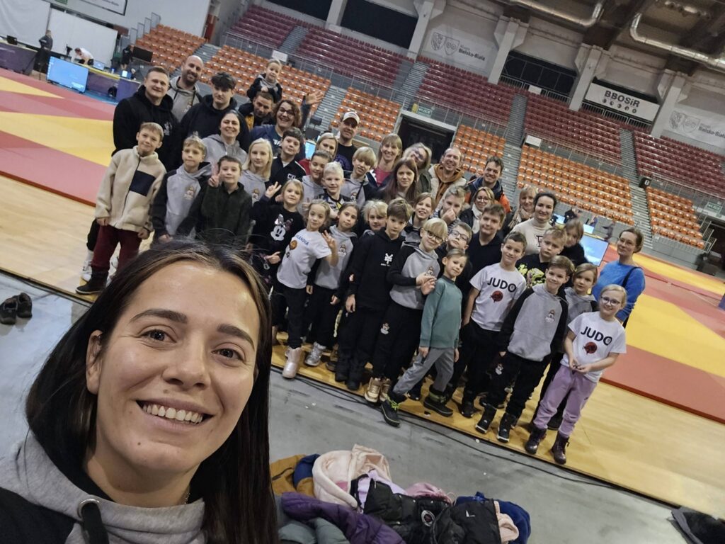 Duża grupa dzieci i dorosłych w bluzach Judo Lion robi wspólne selfie na matach w dużej arenie ze stadionowymi krzesełkami.