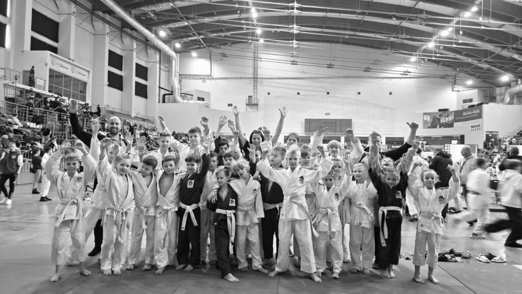 Czarno-białe zdjęcie grupy dzieci w judogach z uniesionymi rękami i uśmiechami na hali sportowej po zawodach.