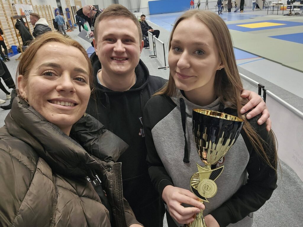 Dwie kobiety i mężczyzna uśmiechają się do selfie w hali, jedna z kobiet trzyma duży czarno-złoty puchar; w tle maty judo.
