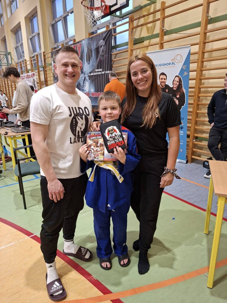 Chłopiec w niebieskim judogu z żółto-białym pasem stoi między trenerami, trzymając folder Grizzly Judo Cup i etui Born to be Judoka.