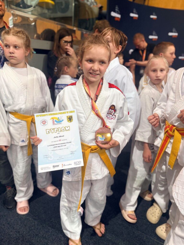 Zawodniczka Judo Lion w białym judogu z żółtym pasem uśmiecha się, trzymając dyplom III miejsca i brązowy medal z Baltic Cup.