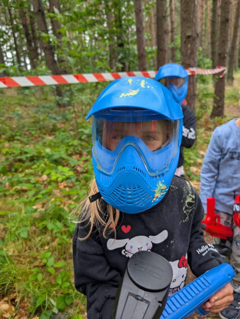 Dziewczynka w niebieskiej masce paintballowej z żółtymi plamami farby trzyma marker w lesie wśród drzew.