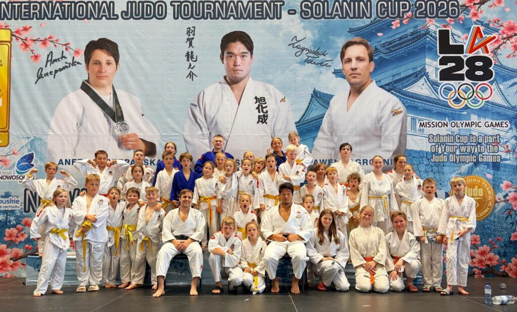 Duża grupa dzieci i trenerów w białych judogach pozuje przed banerem International Judo Tournament Solanin Cup 2026.