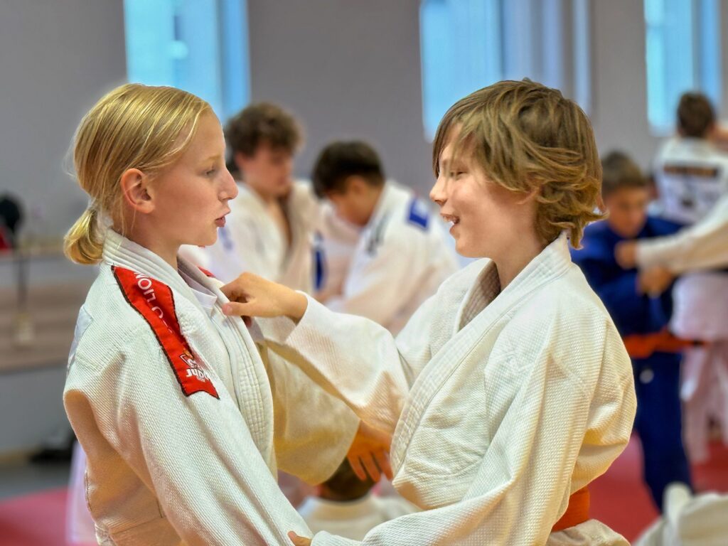 Dwoje dzieci w białych kimonach Judo Lion ćwiczy chwyt na macie i uśmiecha się do siebie