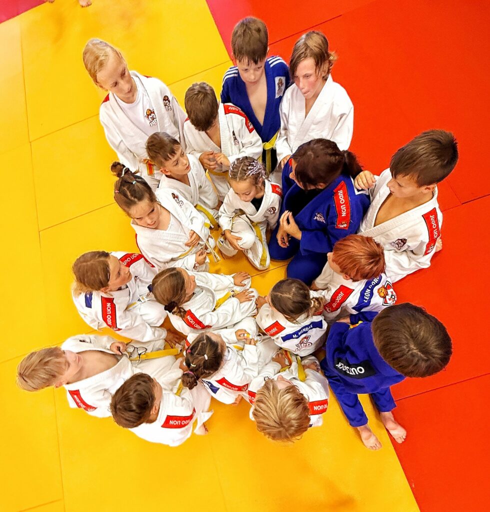 Dzieci i trenerka w kimonach Judo Lion siedzą w kręgu na żółto-czerwonej macie, widok z góry