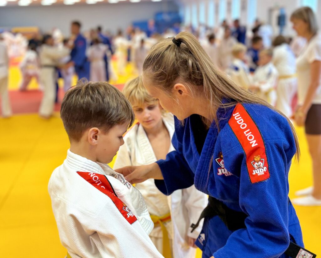 Zawodniczka w niebieskim kimono Judo Lion poprawia strój młodszemu zawodnikowi na macie