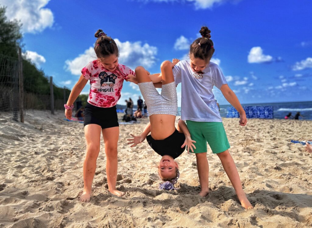 Trzy dziewczynki na plaży, dwie trzymają koleżankę do góry nogami podczas obozu judo w Cetniewie