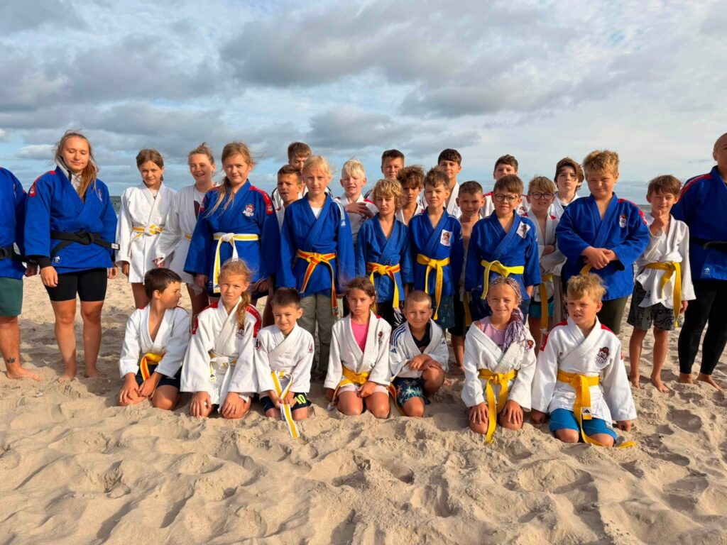Grupa zawodników Judo Lion w białych i niebieskich kimonach pozuje na plaży w Jarosławcu